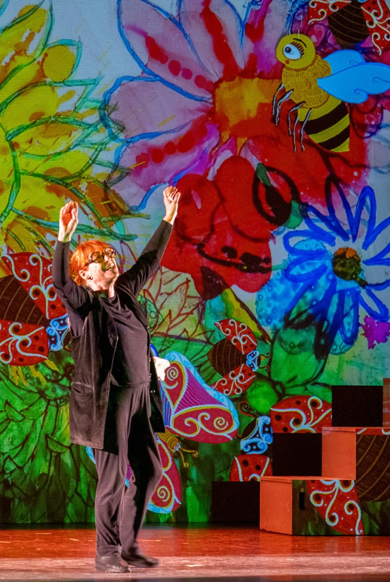 IN UNA COLORATISSIMA SCENOGRAFIA L'ATTRICE RECITA A BRACCIA ALZATE