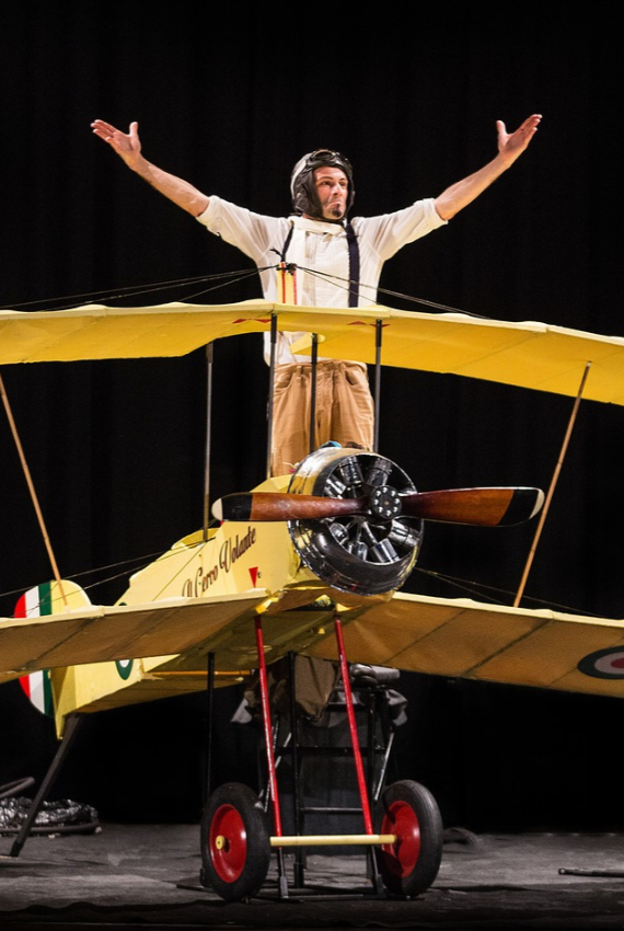 SUL SUO GIGANTESCO BIPLANO GIALLO, L'ATTORE ALZA LE BRACCIA SUL PALCO, NEI PANNI DELL'AVIATORE