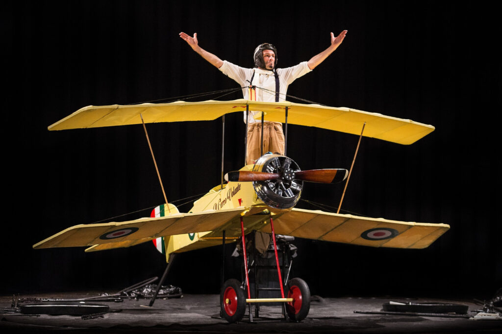 SUL SUO GIGANTESCO BIPLANO GIALLO, L'ATTORE ALZA LE BRACCIA SUL PALCO, NEI PANNI DELL'AVIATORE