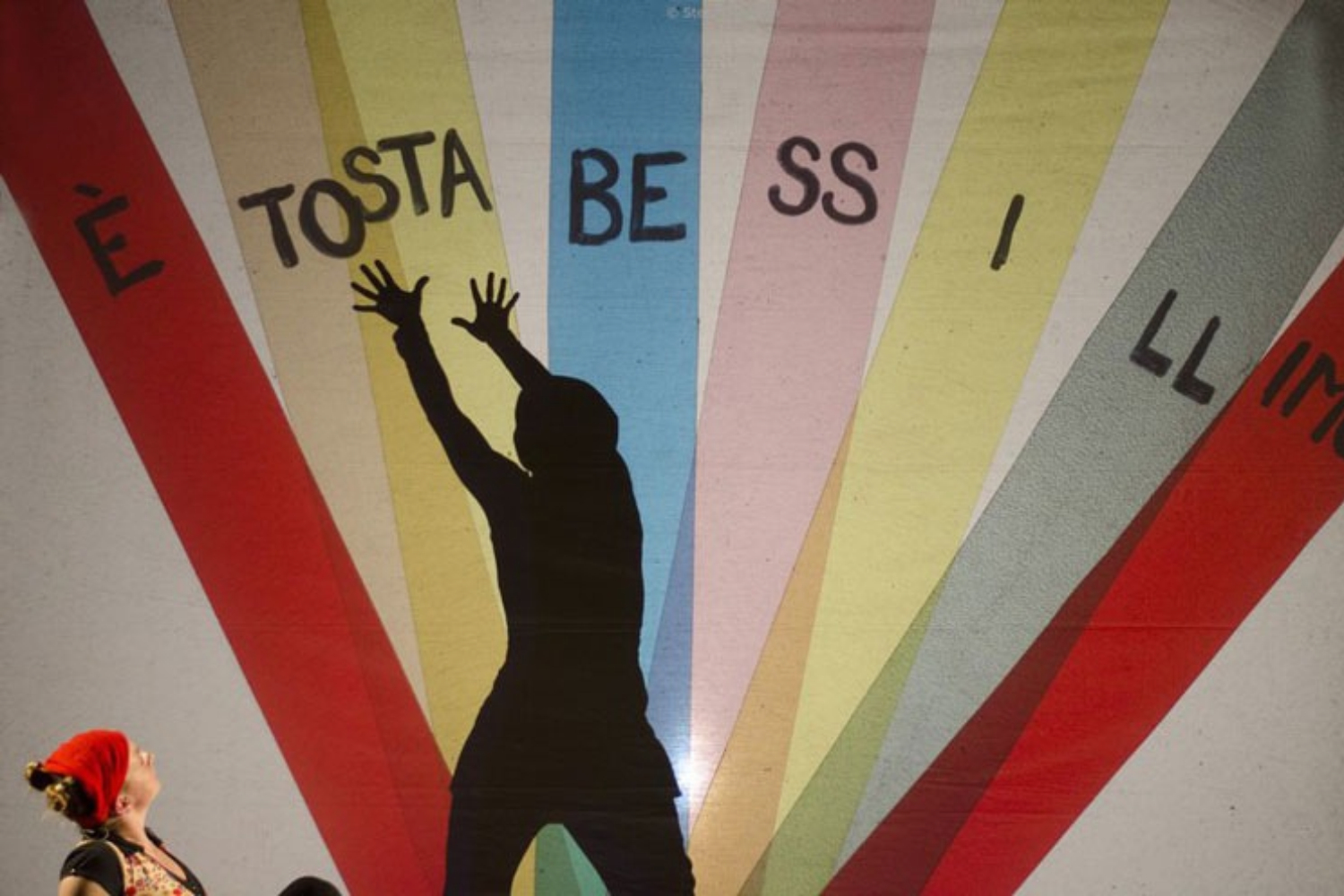 una sagoma umana allunga le mani verso un murales a strisce colorate