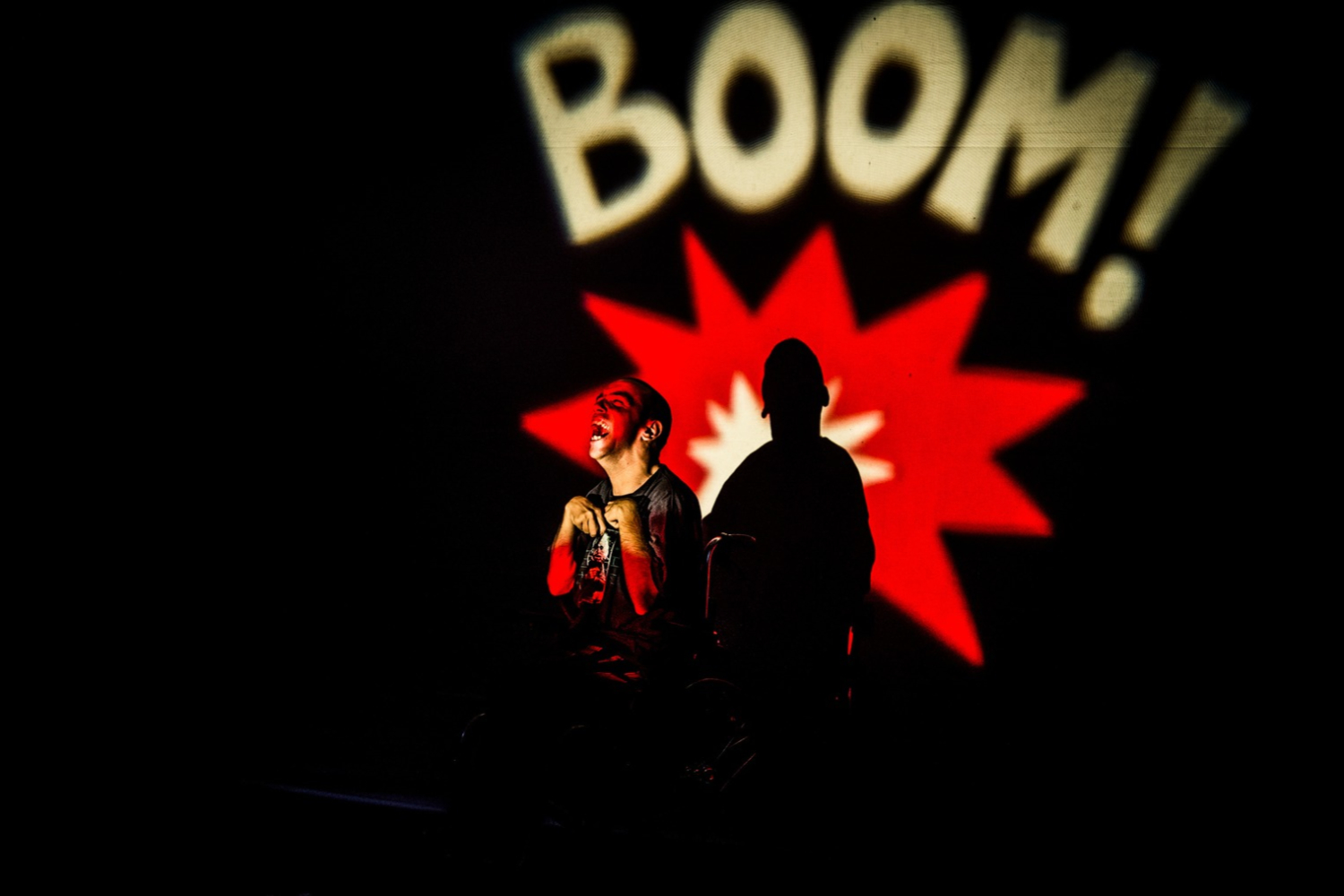 uno dei ragazzi protagonisti in scena davanti a una luminosa scritta proiettata syllo sfondo che recita BOOM